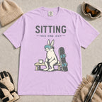 Orchid tee featuring a bunny in ski goggles sitting on a bench beside a snowboard and hot drink with the phrase “Sitting This One Out.”