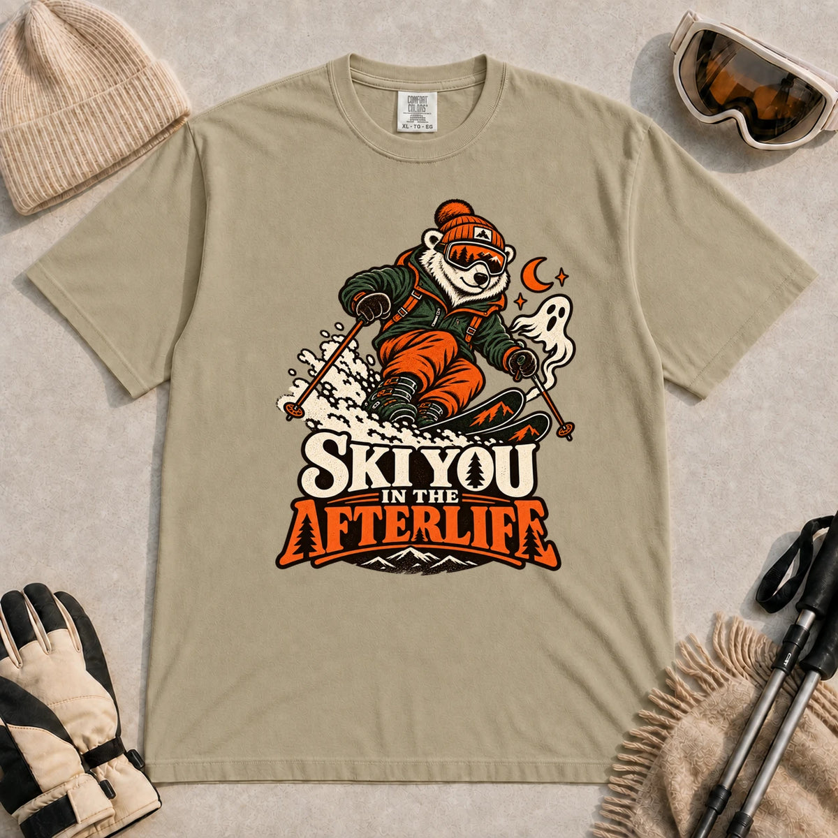 Khaki tee featuring a polar bear skiing through a spooky Halloween scene with a ghost and crescent moon above the phrase “Ski You in the Afterlife.”