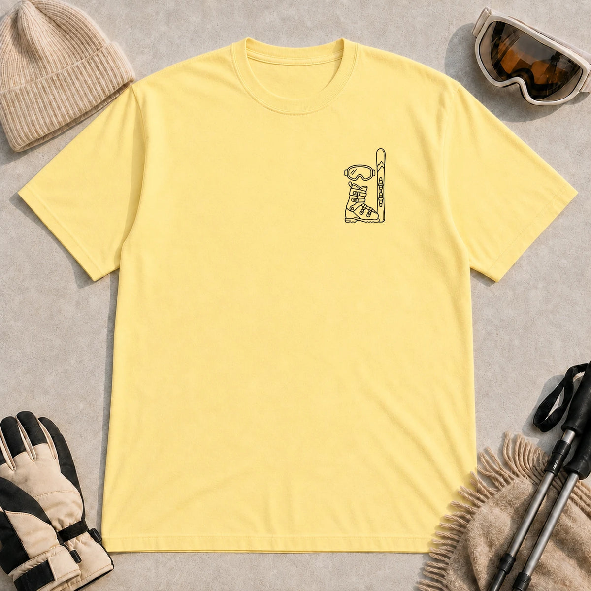 Cornsilk tee featuring minimalist black line art of ski goggles, a ski boot, and an upright ski on the left chest.
