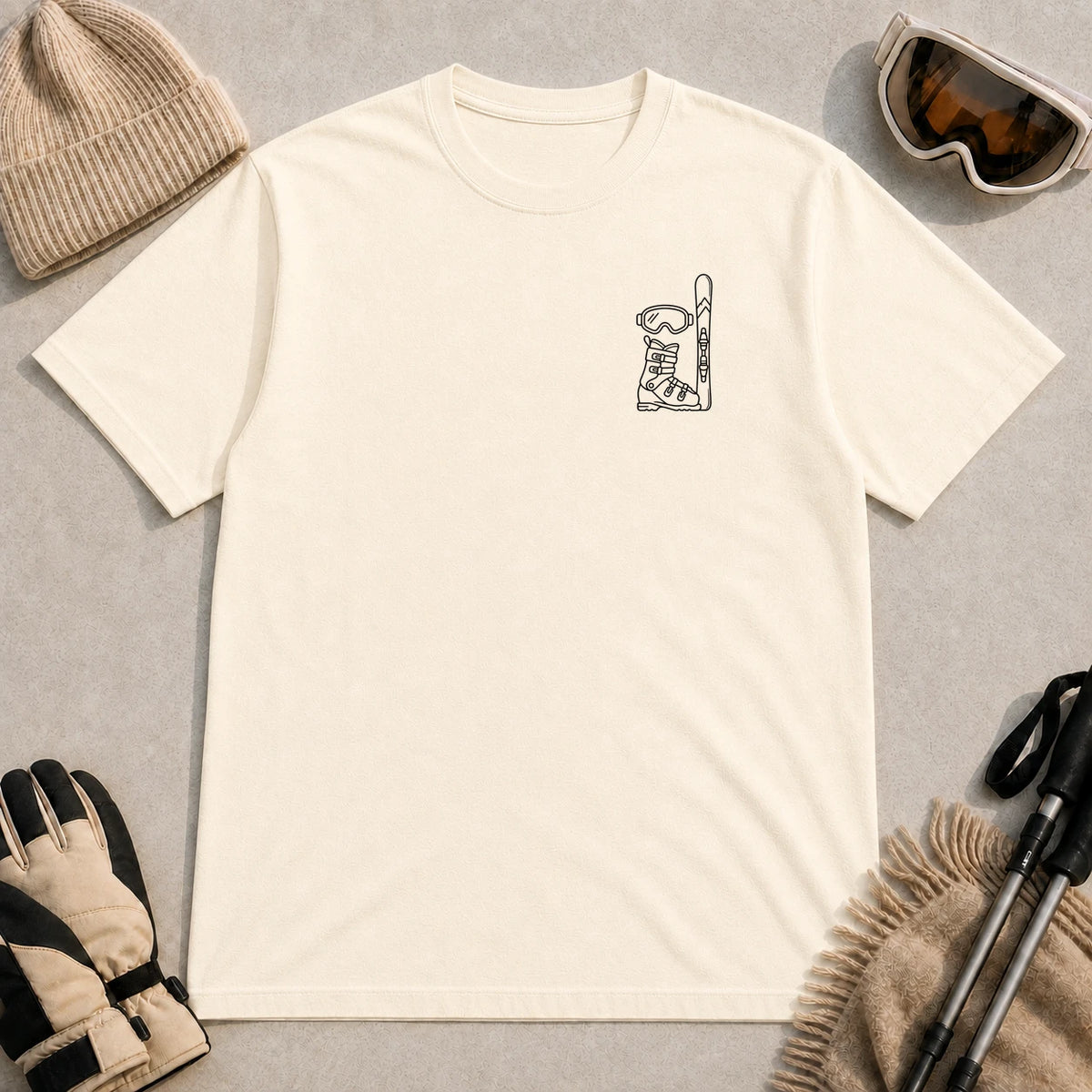 Natural tee featuring minimalist black line art of ski goggles, a ski boot, and an upright ski on the left chest.