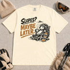 Ivory tee featuring a relaxed porcupine in ski goggles lounging in a chair with a hot drink beside a snowboard, with the phrase “Slopes? Maybe Later.”