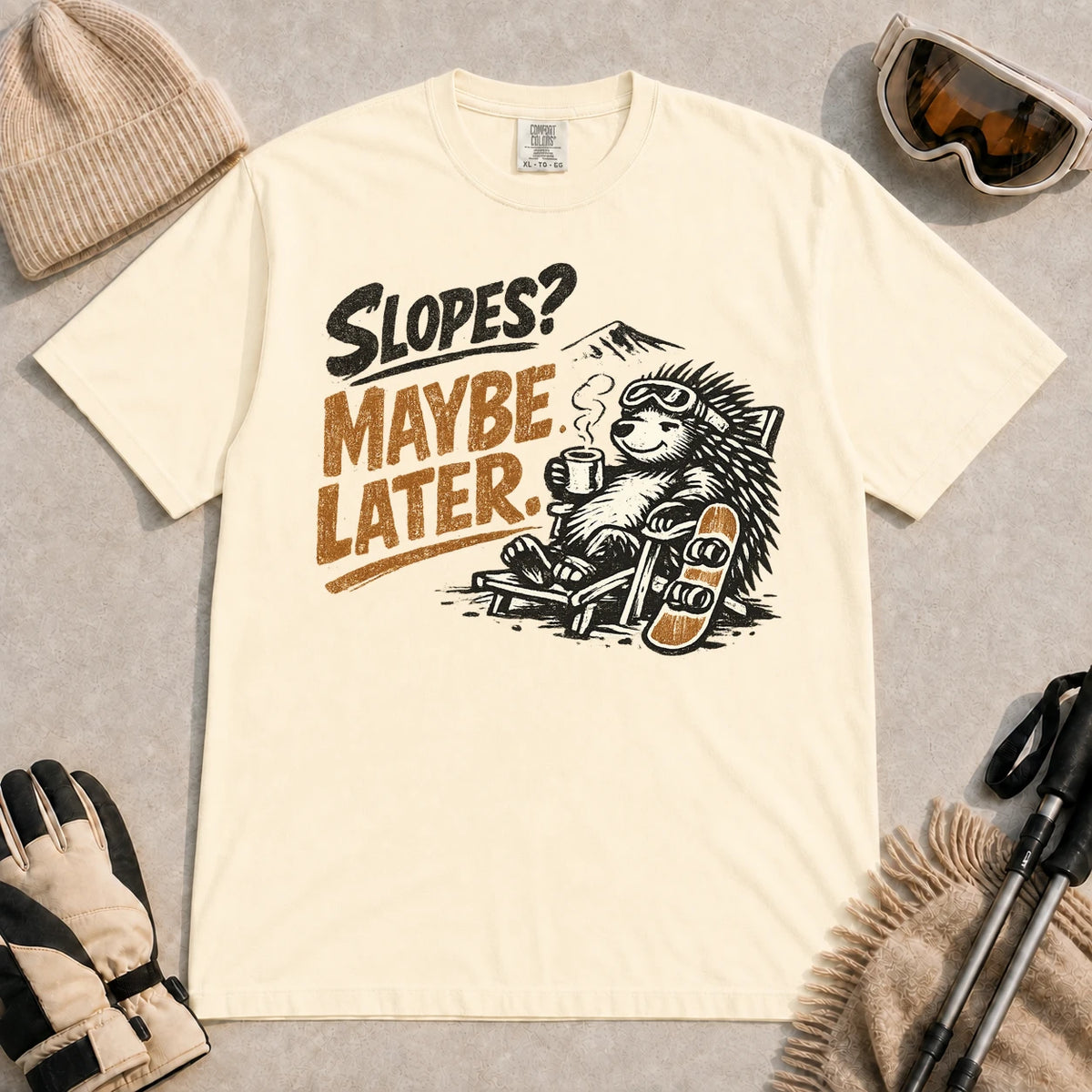 Ivory tee featuring a relaxed porcupine in ski goggles lounging in a chair with a hot drink beside a snowboard, with the phrase “Slopes? Maybe Later.”