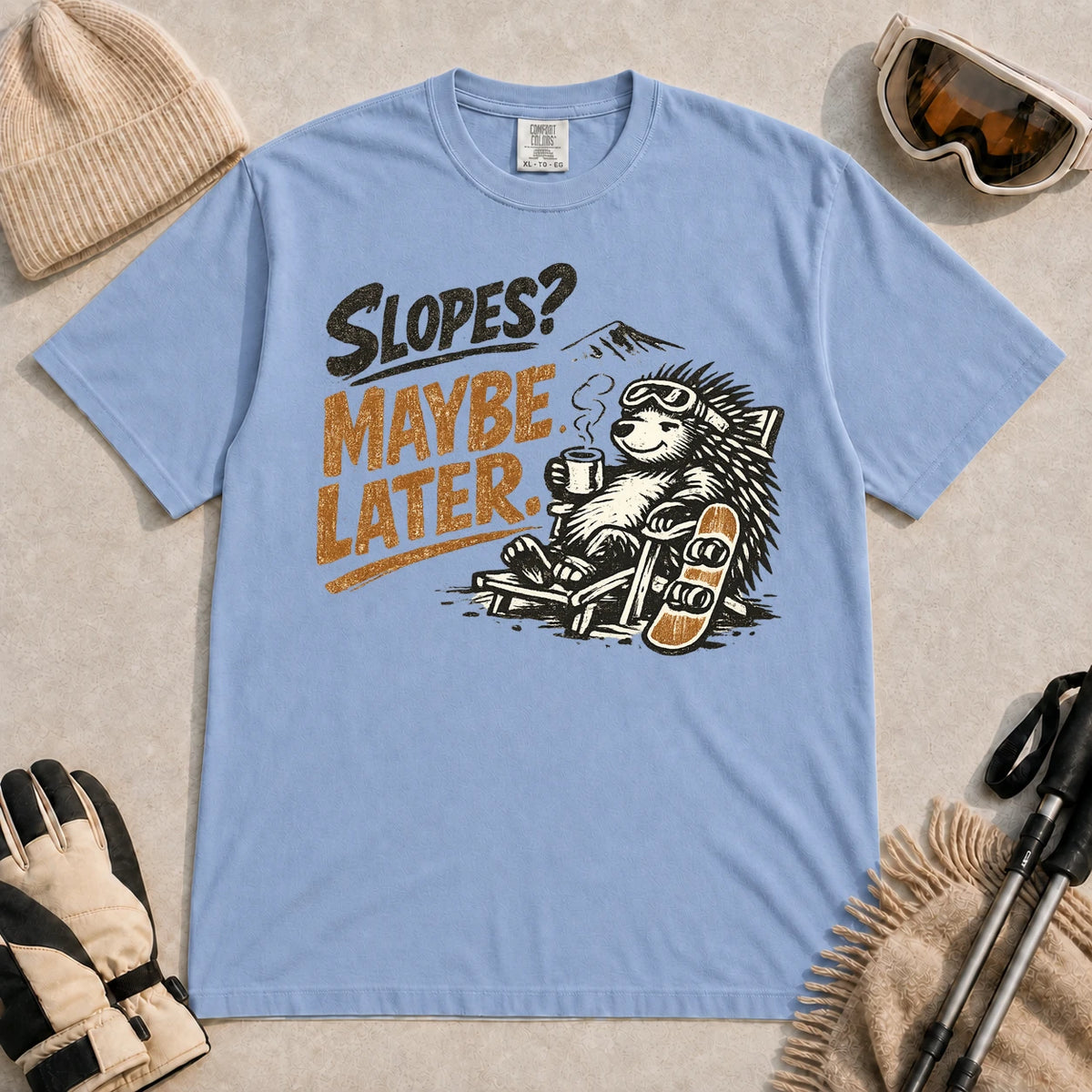 Washed Denim tee featuring a relaxed porcupine in ski goggles lounging in a chair with a hot drink beside a snowboard, with the phrase “Slopes? Maybe Later.”
