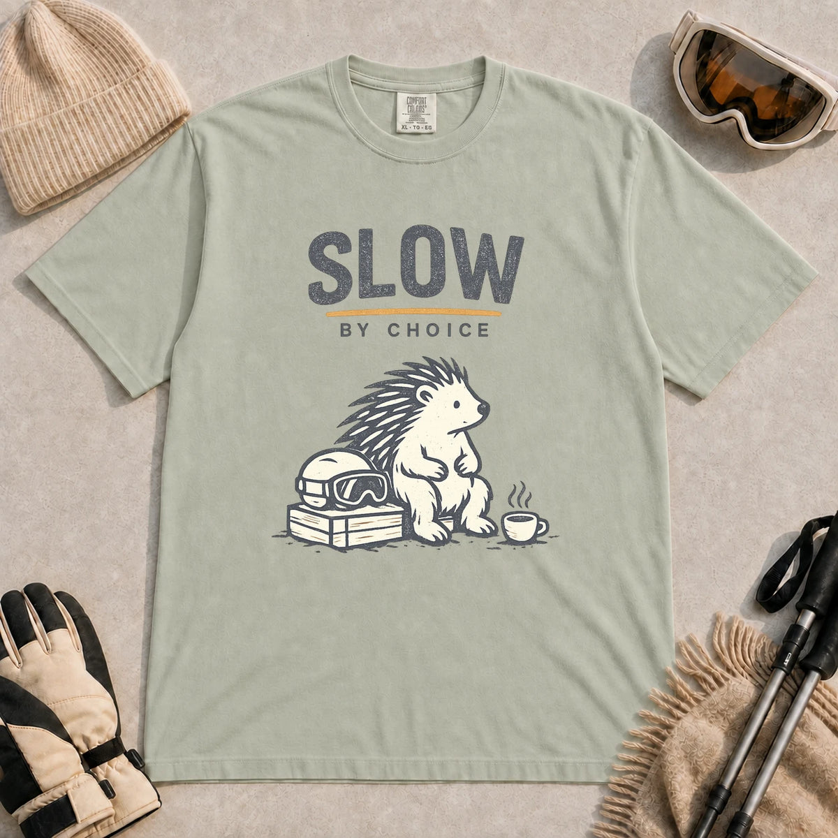 Bay tee featuring a relaxed hedgehog sitting beside ski goggles and a hot drink with the phrase “Slow by Choice.”