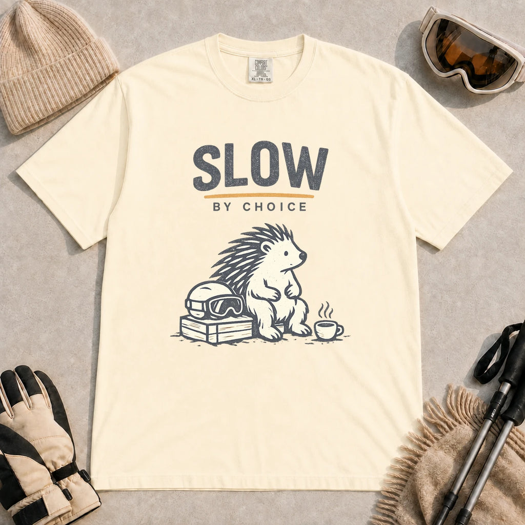 Ivory tee featuring a relaxed hedgehog sitting beside ski goggles and a hot drink with the phrase “Slow by Choice.”