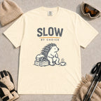 Ivory tee featuring a relaxed hedgehog sitting beside ski goggles and a hot drink with the phrase “Slow by Choice.”