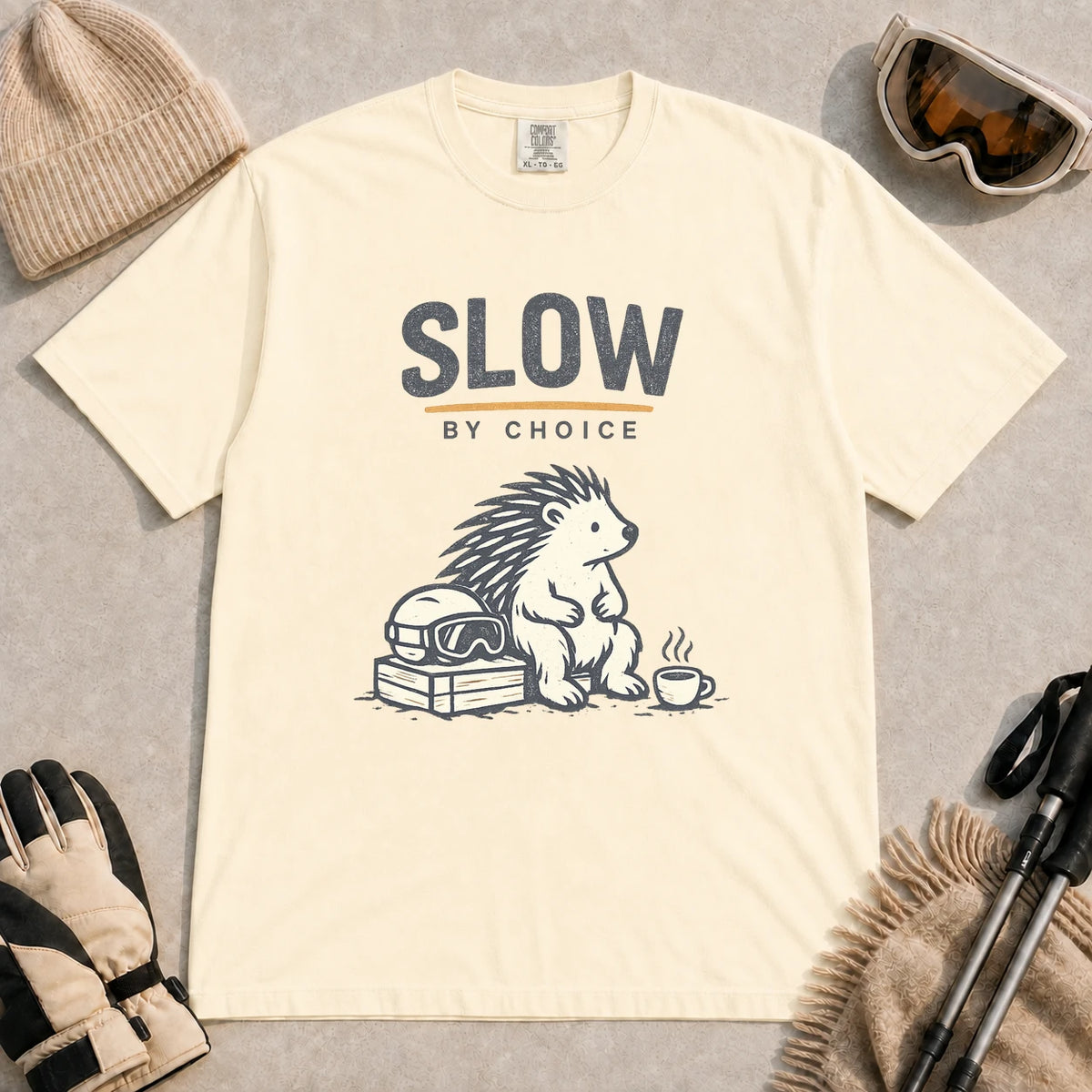 Ivory tee featuring a relaxed hedgehog sitting beside ski goggles and a hot drink with the phrase “Slow by Choice.”