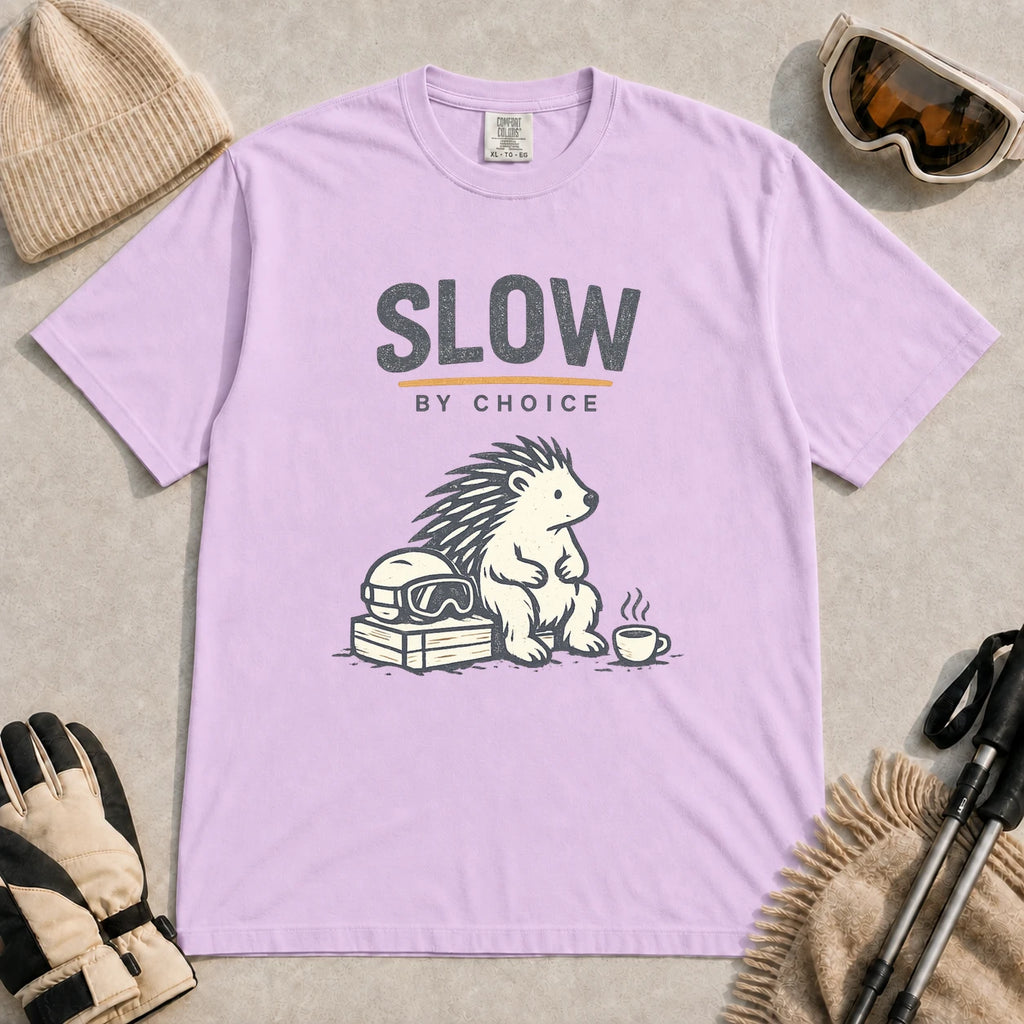 Orchid tee featuring a relaxed hedgehog sitting beside ski goggles and a hot drink with the phrase “Slow by Choice.”