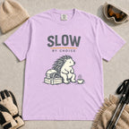 Orchid tee featuring a relaxed hedgehog sitting beside ski goggles and a hot drink with the phrase “Slow by Choice.”