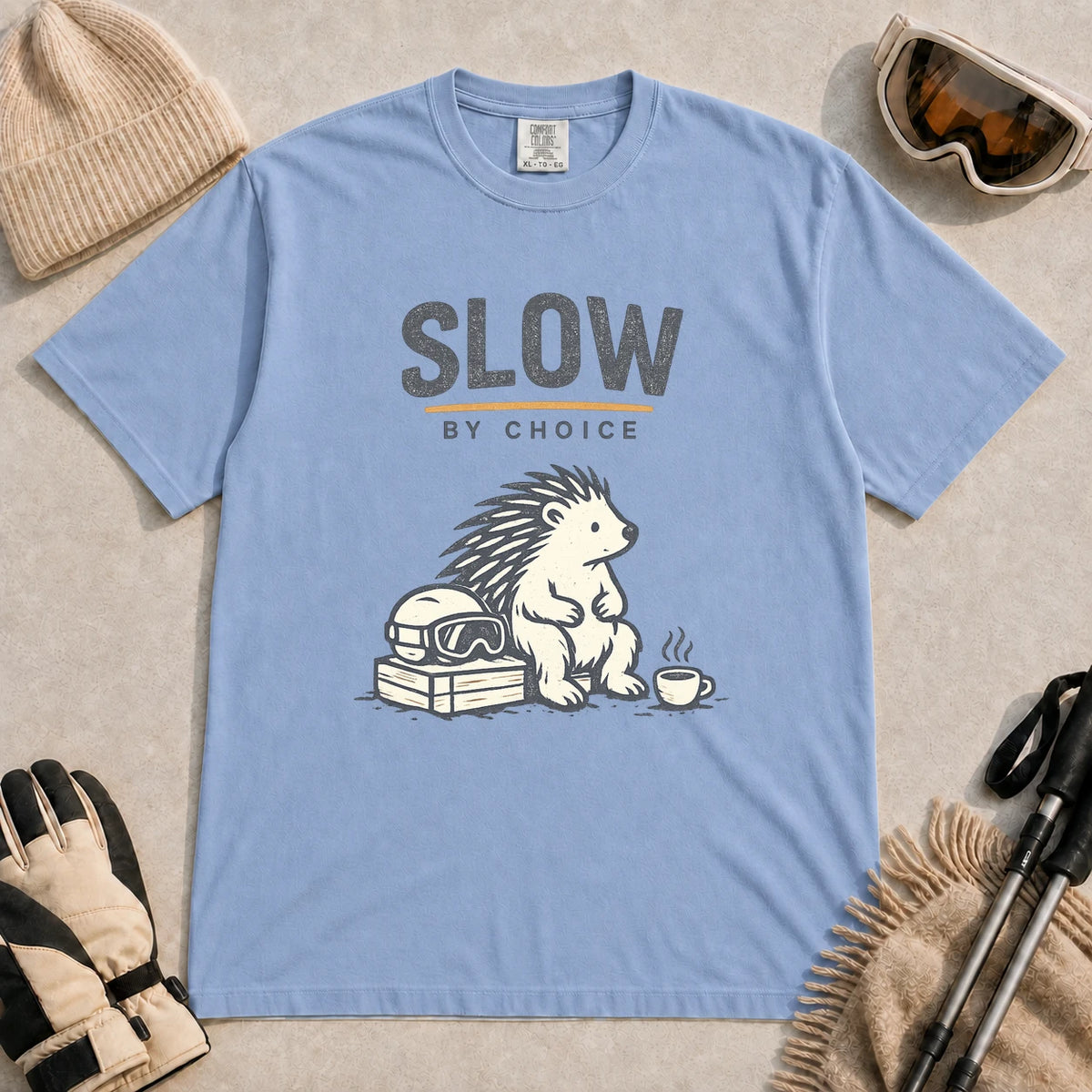 Washed Denim tee featuring a relaxed hedgehog sitting beside ski goggles and a hot drink with the phrase “Slow by Choice.”