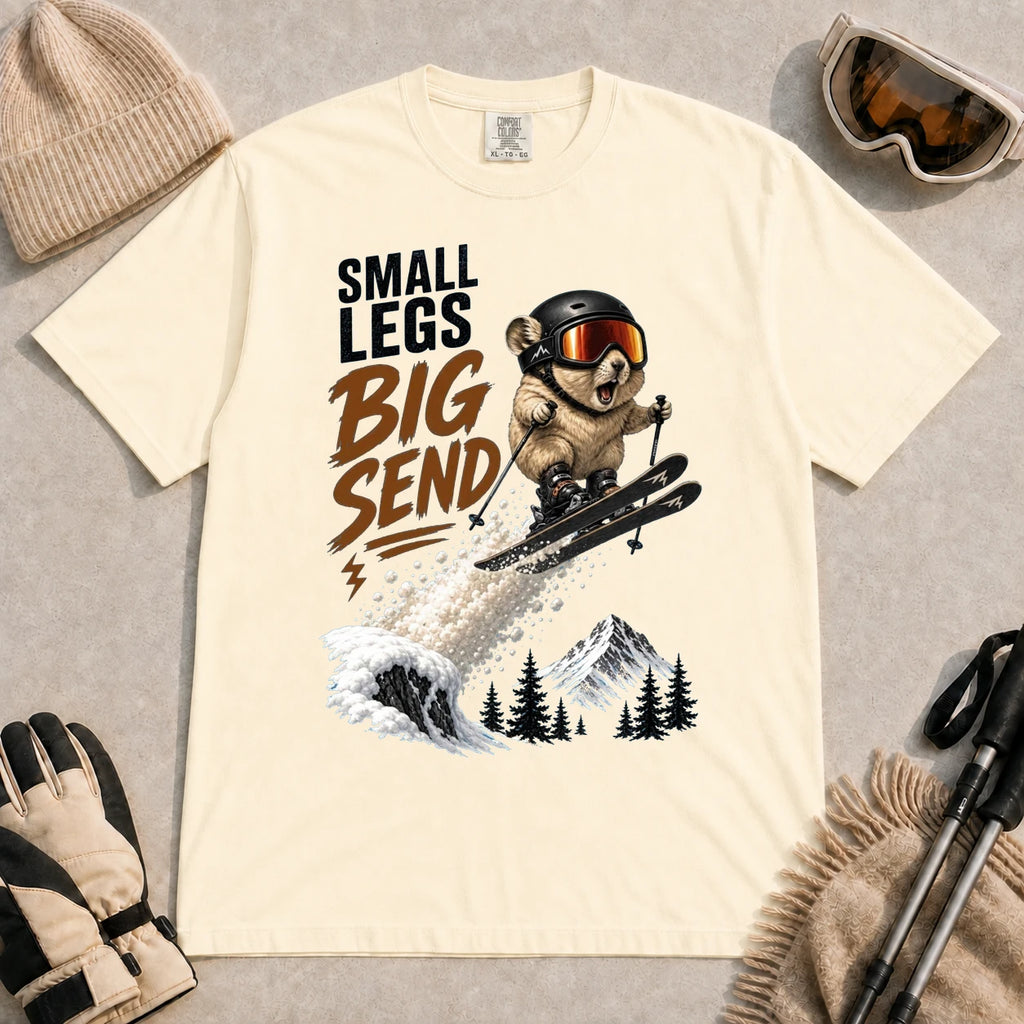 Ivory tee featuring a tiny goggle-wearing animal skier launching off a snowy jump, with mountain scenery and the phrase “Small Legs, Big Send.”