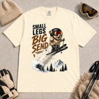 Ivory tee featuring a tiny goggle-wearing animal skier launching off a snowy jump, with mountain scenery and the phrase “Small Legs, Big Send.”