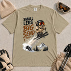 Khaki tee featuring a tiny goggle-wearing animal skier launching off a snowy jump, with mountain scenery and the phrase “Small Legs, Big Send.”