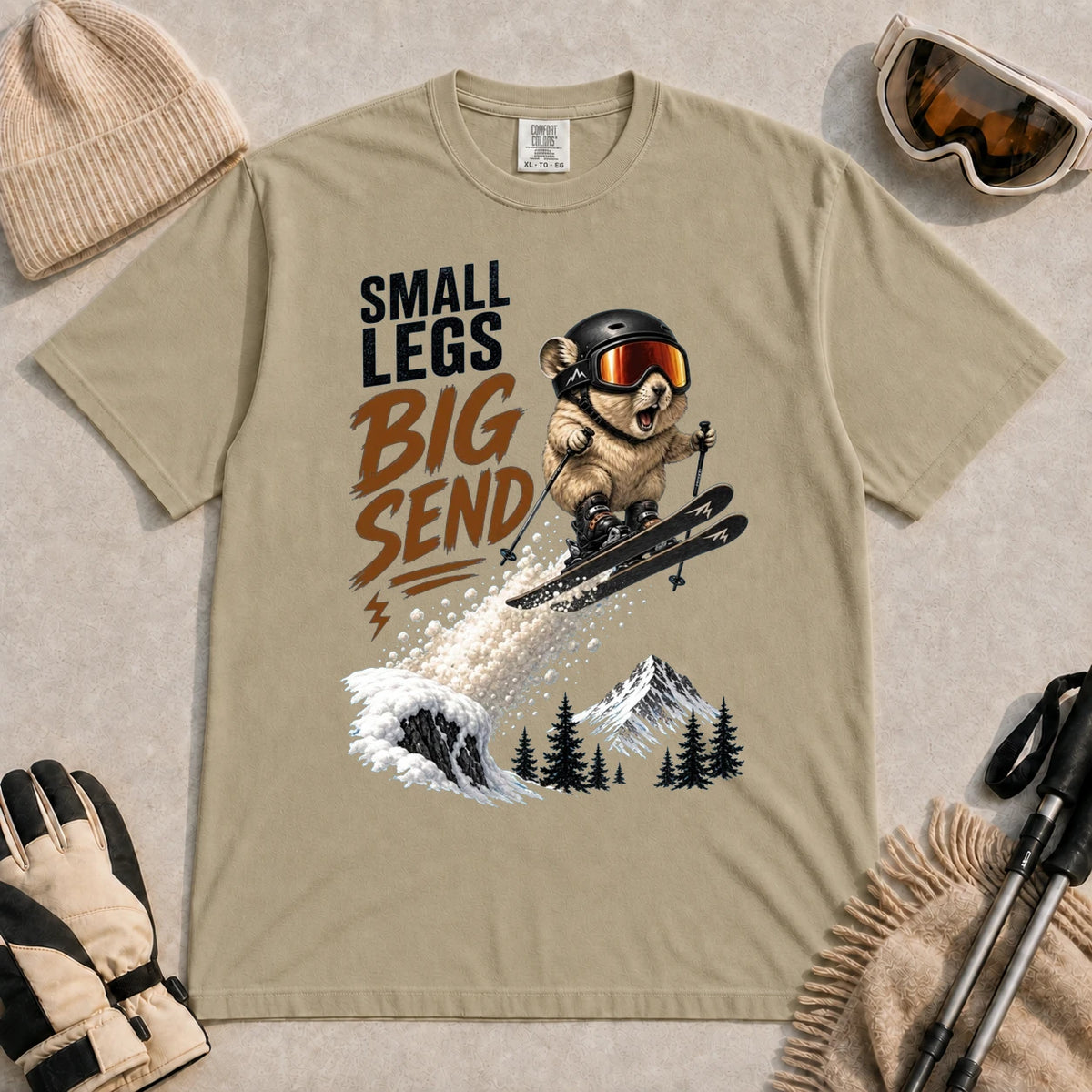 Khaki tee featuring a tiny goggle-wearing animal skier launching off a snowy jump, with mountain scenery and the phrase “Small Legs, Big Send.”