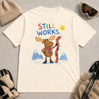 Natural tee featuring a cheerful moose holding a bent red ski in a colorful hand-drawn mountain scene with the phrase “Still Works.”