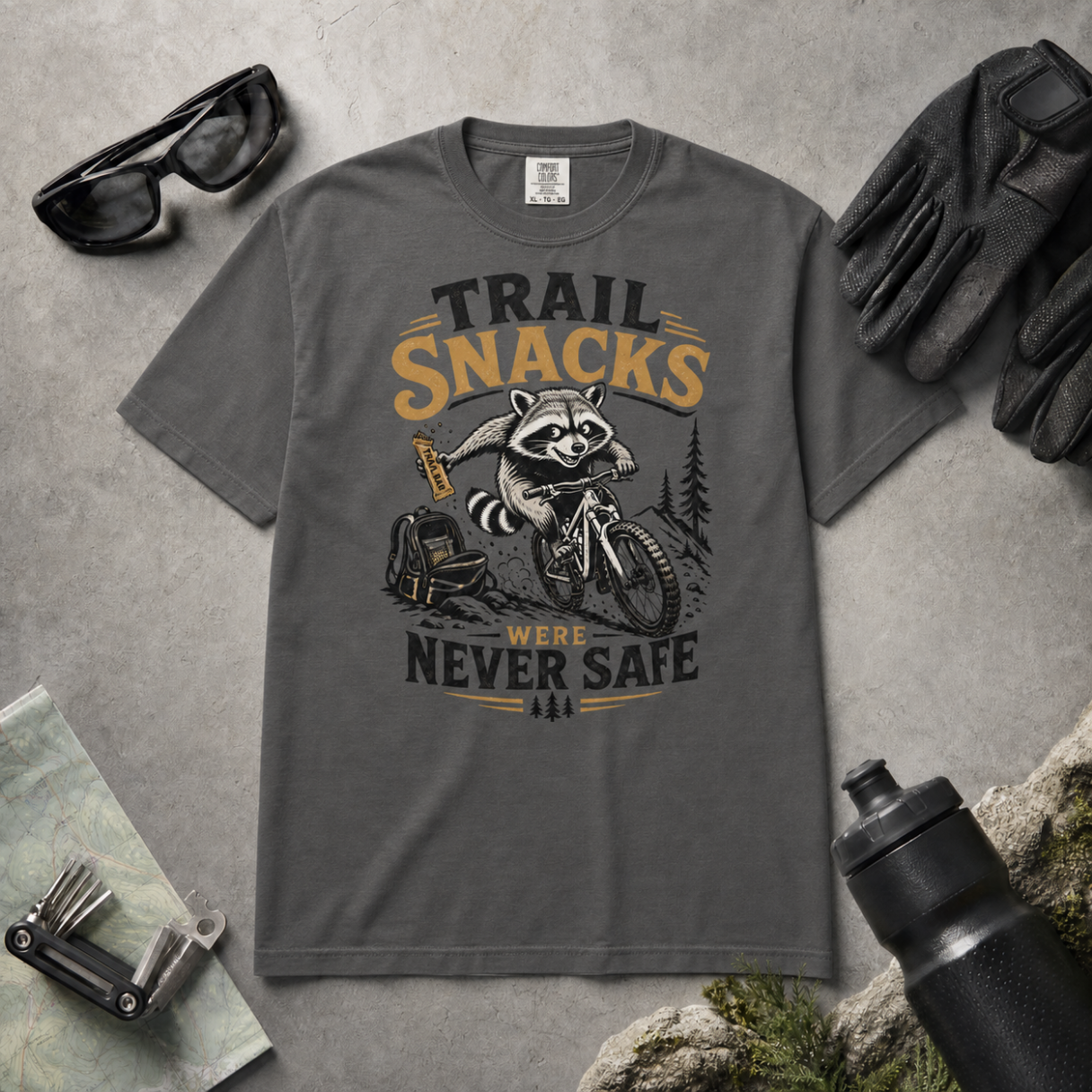 Pepper gray t-shirt with a raccoon graphic and text 'Trail Snacks Were Never Safe' on a gray surface with outdoor accessories.