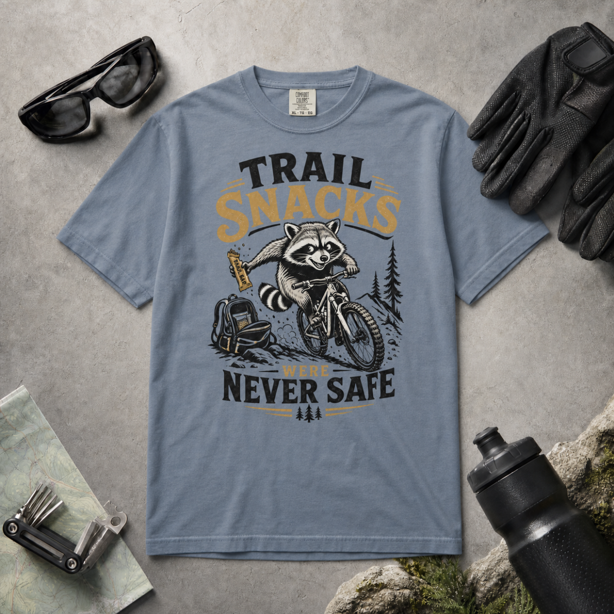 Blue jean colored t-shirt with a raccoon graphic and text on a gray surface with outdoor gear.