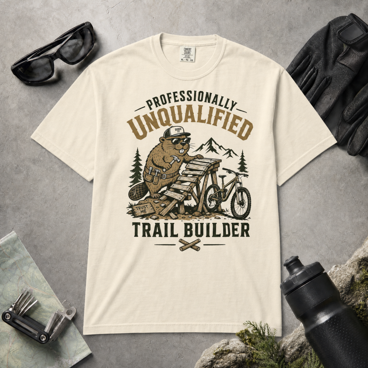 Ivory white t-shirt with a graphic of a beaver building a trail, surrounded by outdoor gear on a gray surface.