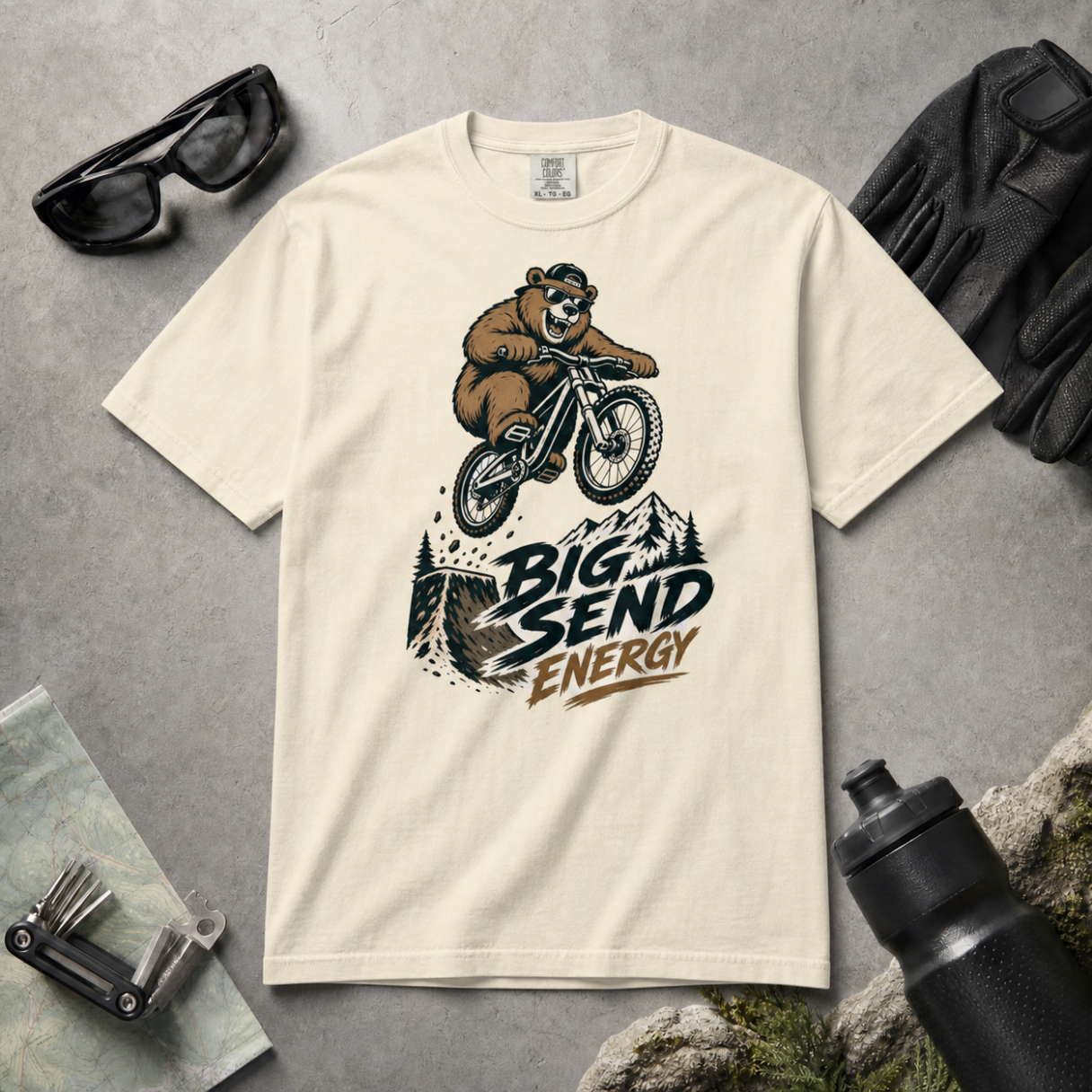 Ivory colored t-shirt with bear riding a bike graphic and 'Big Send Energy' text on a gray surface with accessories.