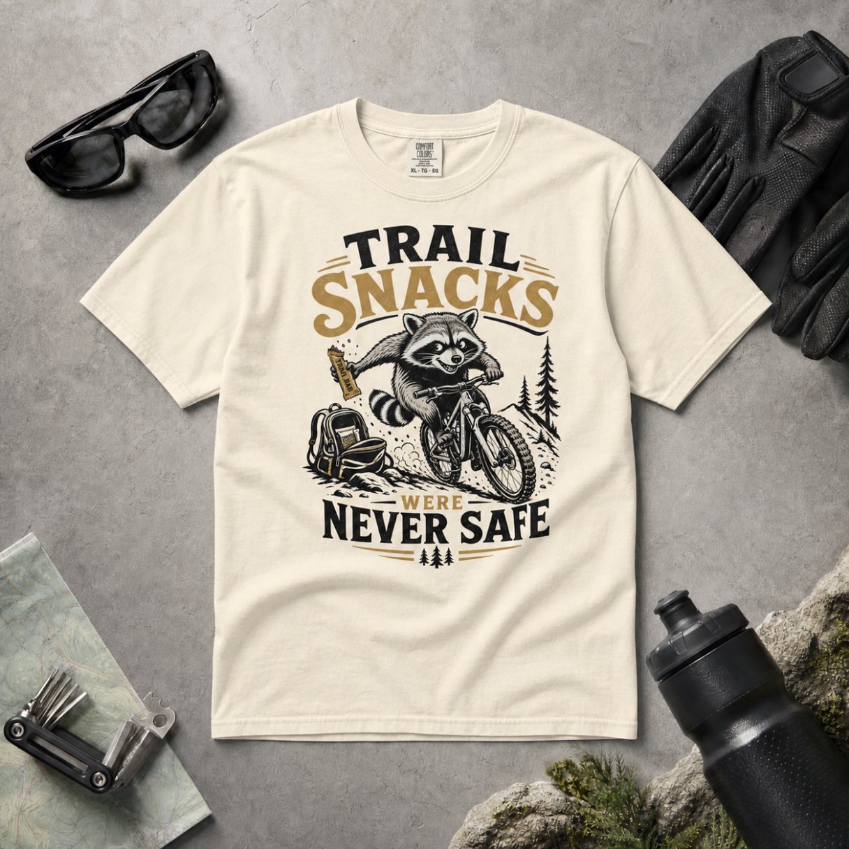 Ivory colored t-shirt with raccoon graphic and text 'Trail Snacks Were Never Safe' on a gray surface with outdoor accessories.