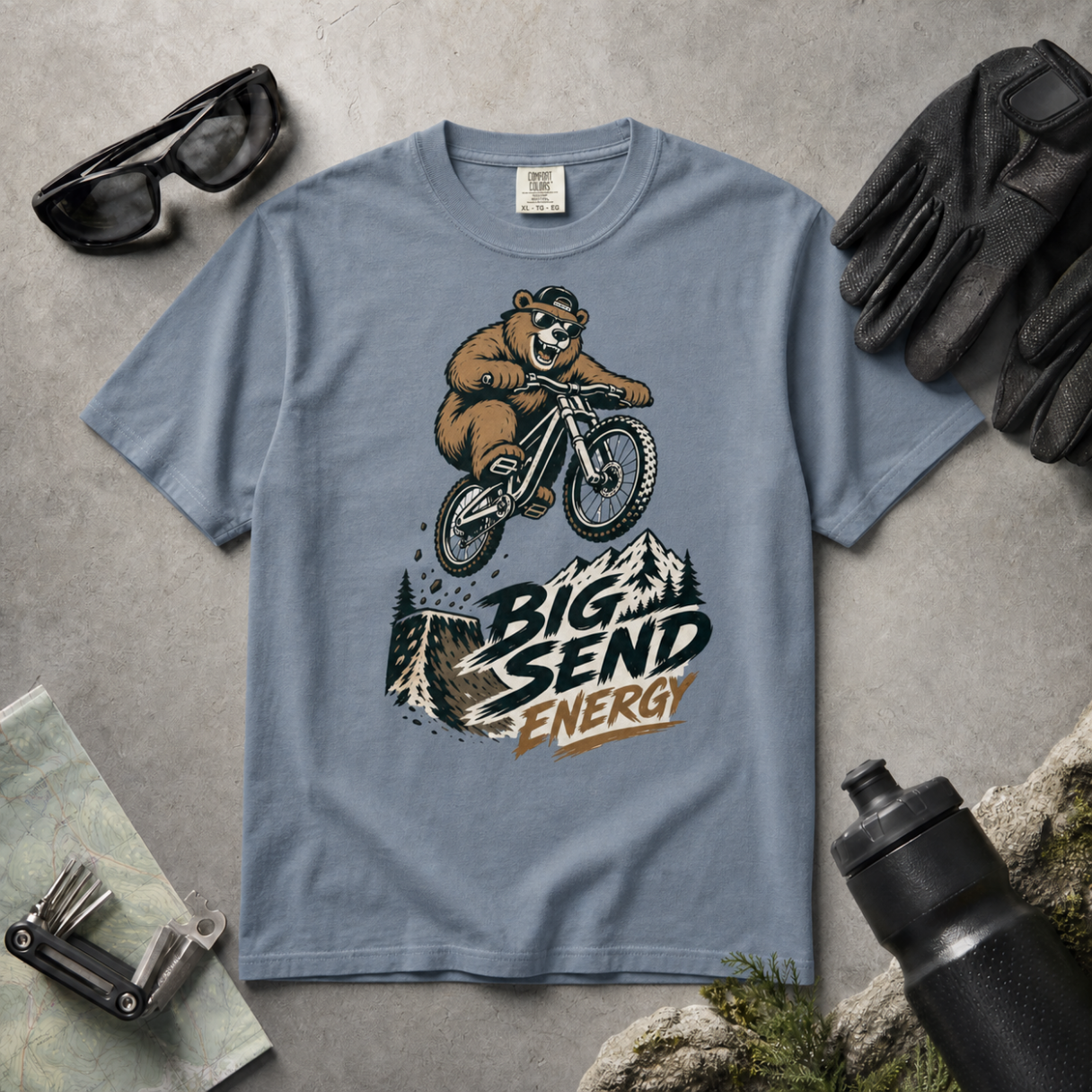 Blue jean colored t-shirt with bear riding a bike graphic and 'Big Send Energy' text, surrounded by sunglasses, gloves, and a water bottle on a concrete surface.