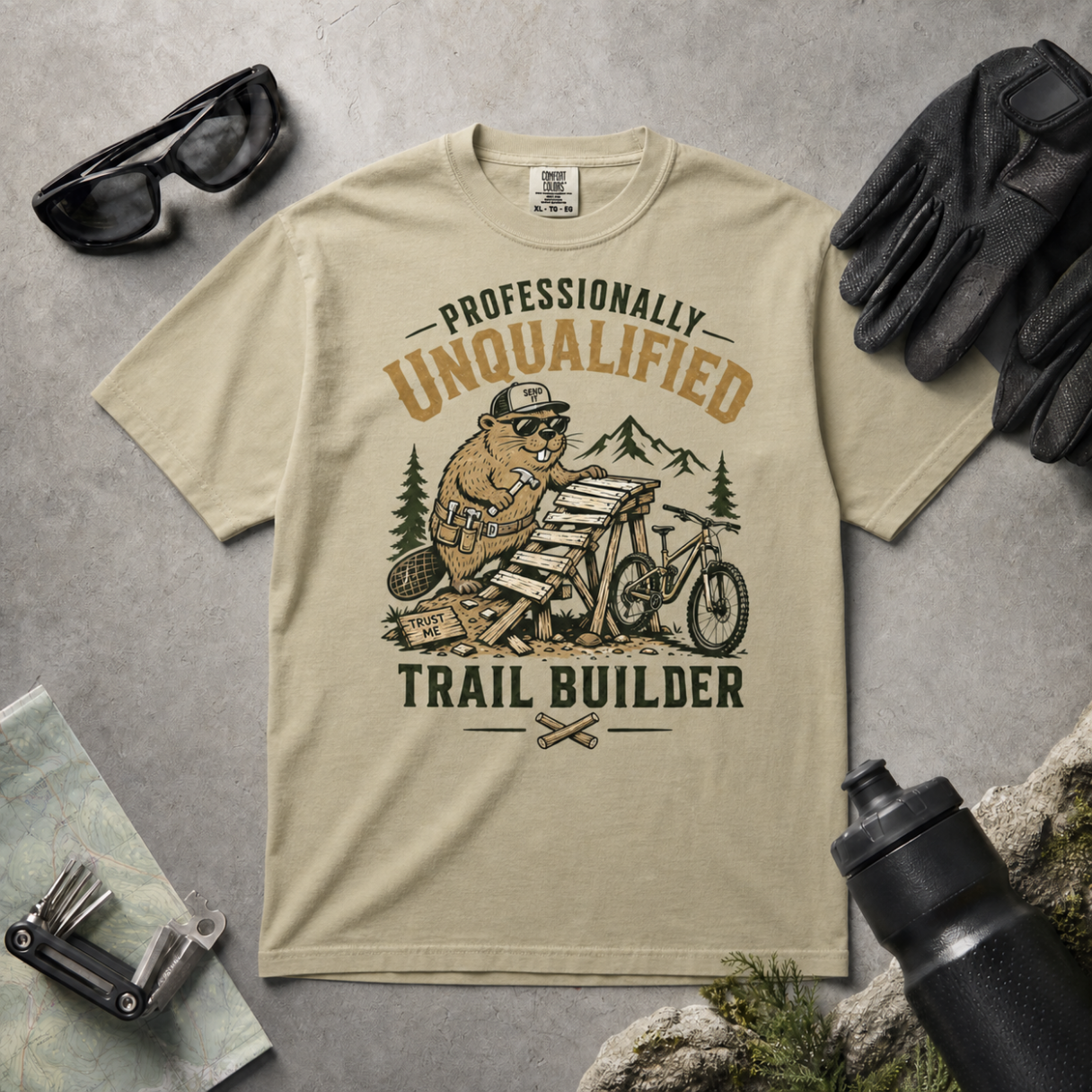 Khaki colored t-shirt with beaver graphic and text 'Professionally Unqualified Trail Builder' on a gray surface with accessories.