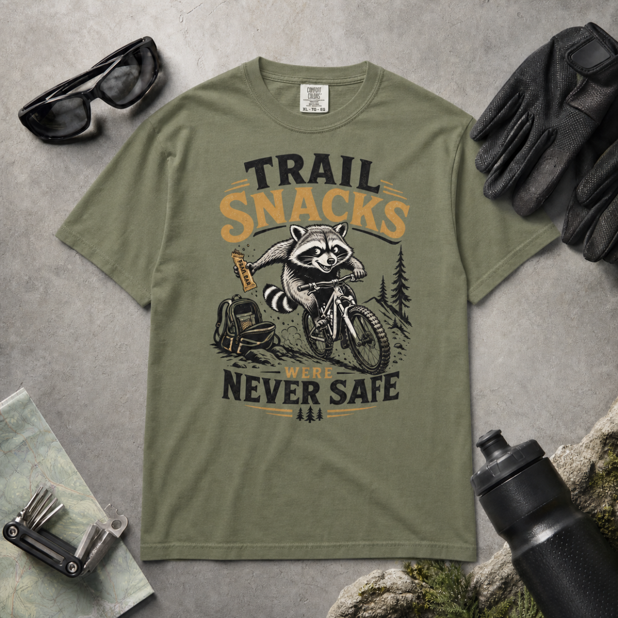 Moss green t-shirt with a raccoon graphic and text on a gray surface with accessories.
