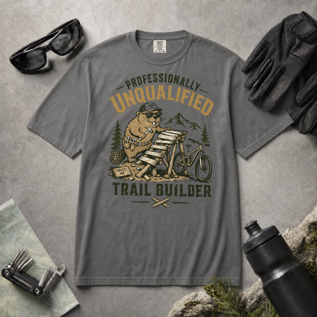 Pepper gray t-shirt with beaver graphic and text 'Professionally Unqualified Trail Builder' on a concrete surface with outdoor gear.
