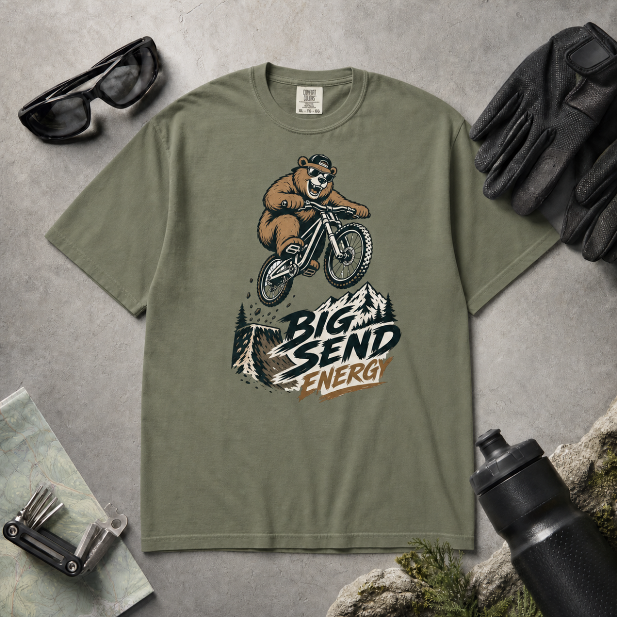 Moss green t-shirt with bear riding a bike graphic and 'Big Send Energy' text, surrounded by sunglasses, gloves, and a water bottle on a gray surface.