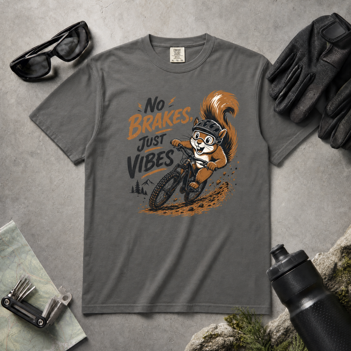 Pepper gray t-shirt with a squirrel graphic and text 'No Brakes Just Vibes' on a gray surface with accessories.