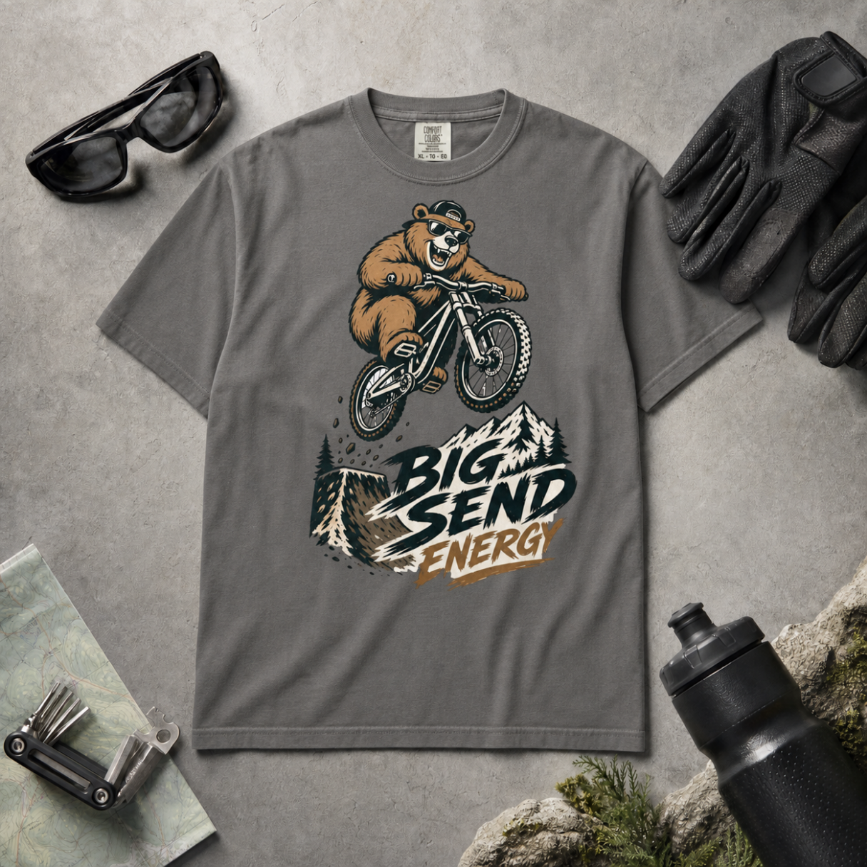 Pepper gray t-shirt with bear riding a bike graphic and 'Big Send Energy' text, surrounded by sunglasses, gloves, and a water bottle on a concrete surface.