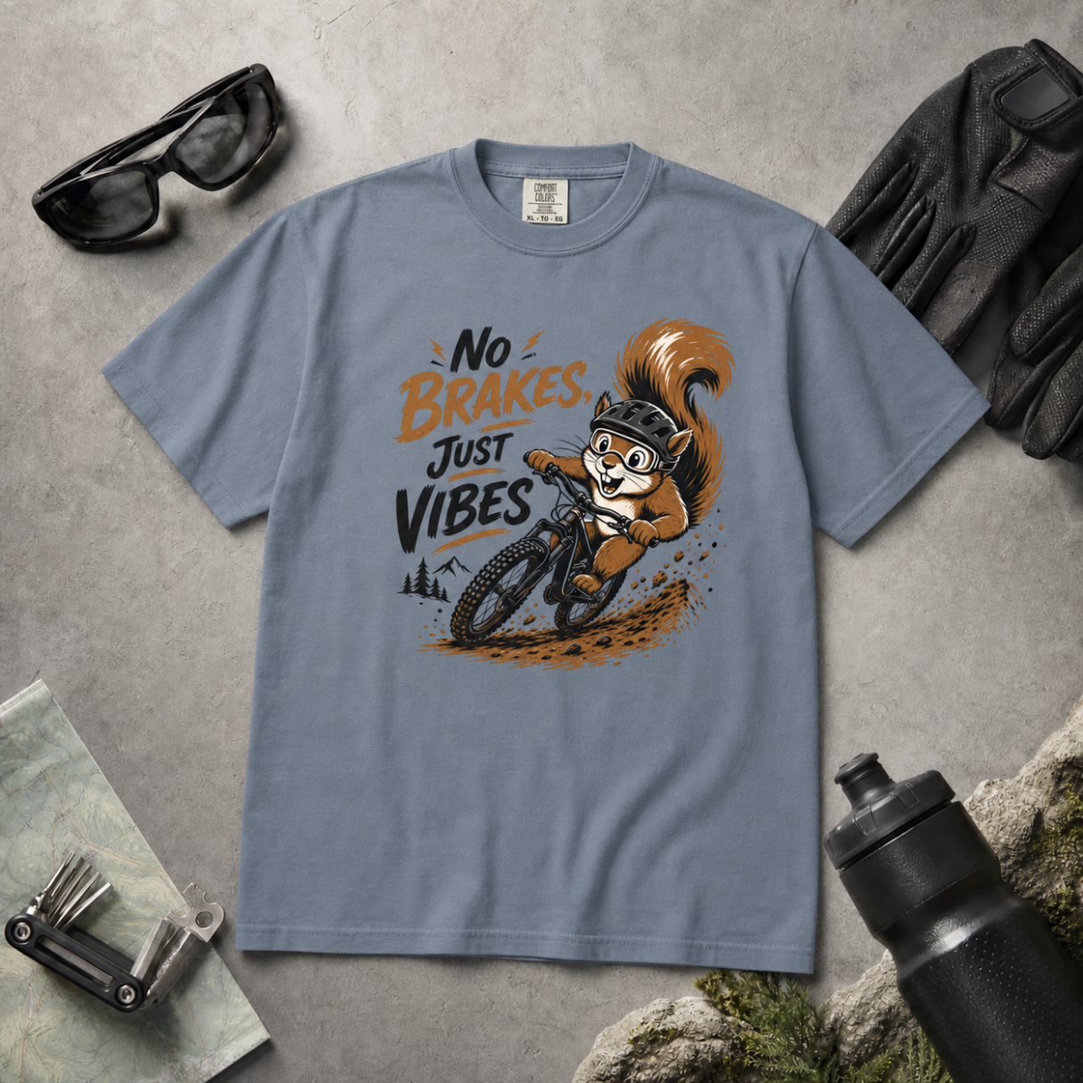Blue jean colored t-shirt with a squirrel graphic and text 'No Brakes Just Vibes' on a gray surface with accessories.