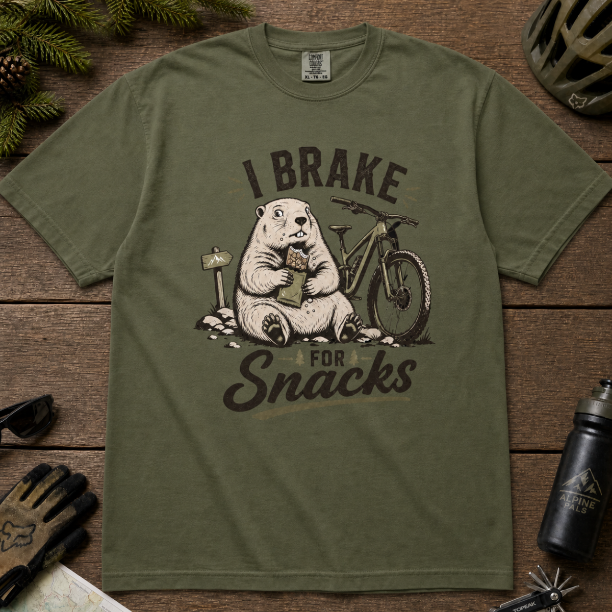 Flat lay of a hemp green t-shirt featuring a playful “I Brake for Snacks” mountain biking design with a marmot eating a snack beside a bike.