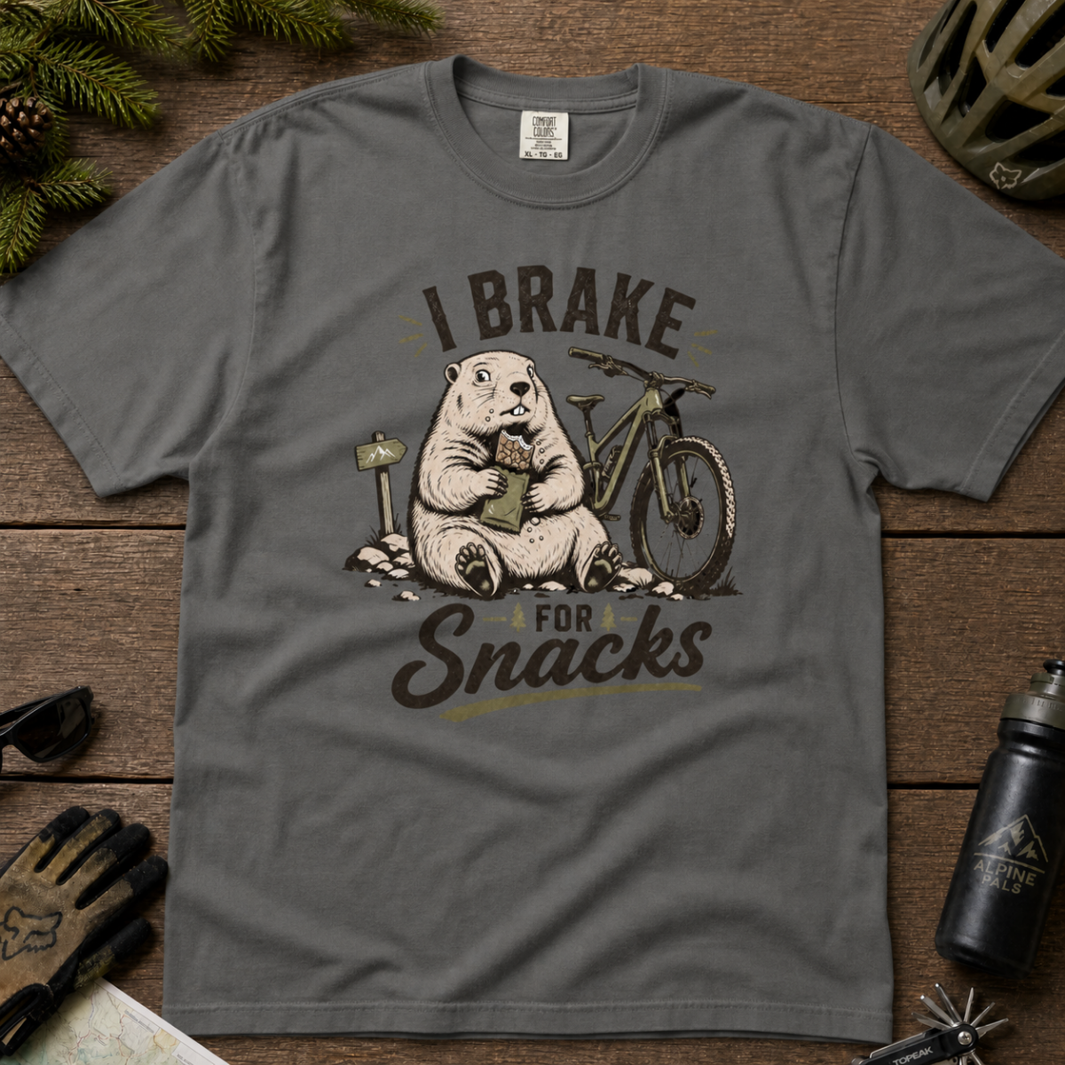 Flat lay of a pepper gray t-shirt featuring a playful “I Brake for Snacks” mountain biking design with a marmot eating a snack beside a bike.