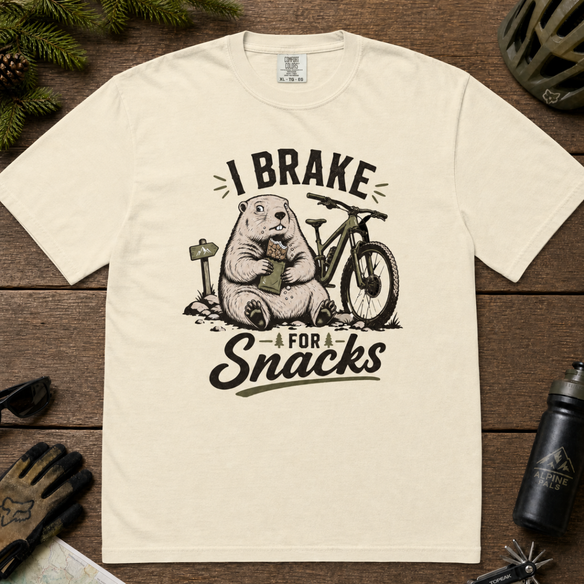 Flat lay of an ivory white t-shirt featuring a playful “I Brake for Snacks” mountain biking design with a marmot eating a snack beside a bike.