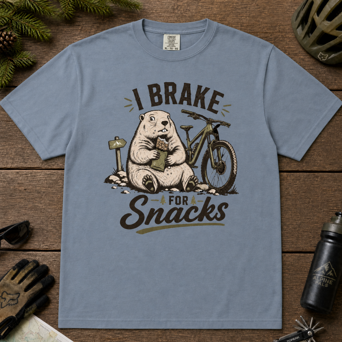 Flat lay of a blue jean colored t-shirt featuring a playful “I Brake for Snacks” mountain biking design with a marmot eating a snack beside a bike.