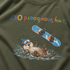 Close-up of a hemp green t-shirt showing a crashed otter in ski goggles with a snowboard flying overhead and colourful upside-down “Is My Snowboard OK?” text.