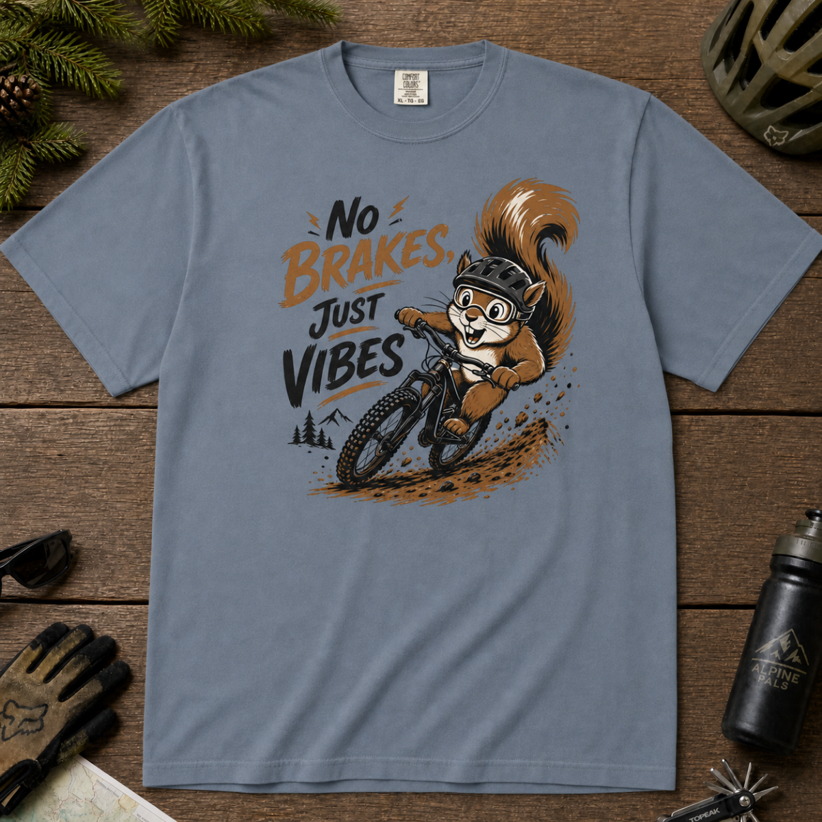 Blue jean colored t-shirt featuring a squirrel mountain biking down a dirt trail with bold “No Brakes, Just Vibes” lettering.