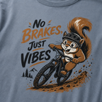 Close-up of a Blue jean colored t-shirt featuring a helmeted squirrel riding a mountain bike with “No Brakes, Just Vibes” text.