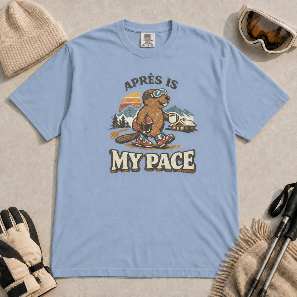Washed Denim t-shirt featuring an “Après Is My Pace” design with a beaver wearing ski goggles and boots, carrying a helmet and hot drink against a retro mountain and chalet scene.