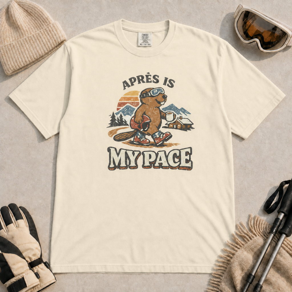 Ivory t-shirt featuring an “Après Is My Pace” design with a beaver wearing ski goggles and boots, carrying a helmet and hot drink against a retro mountain and chalet scene.