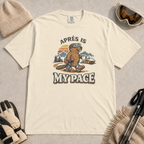 Ivory t-shirt featuring an “Après Is My Pace” design with a beaver wearing ski goggles and boots, carrying a helmet and hot drink against a retro mountain and chalet scene.