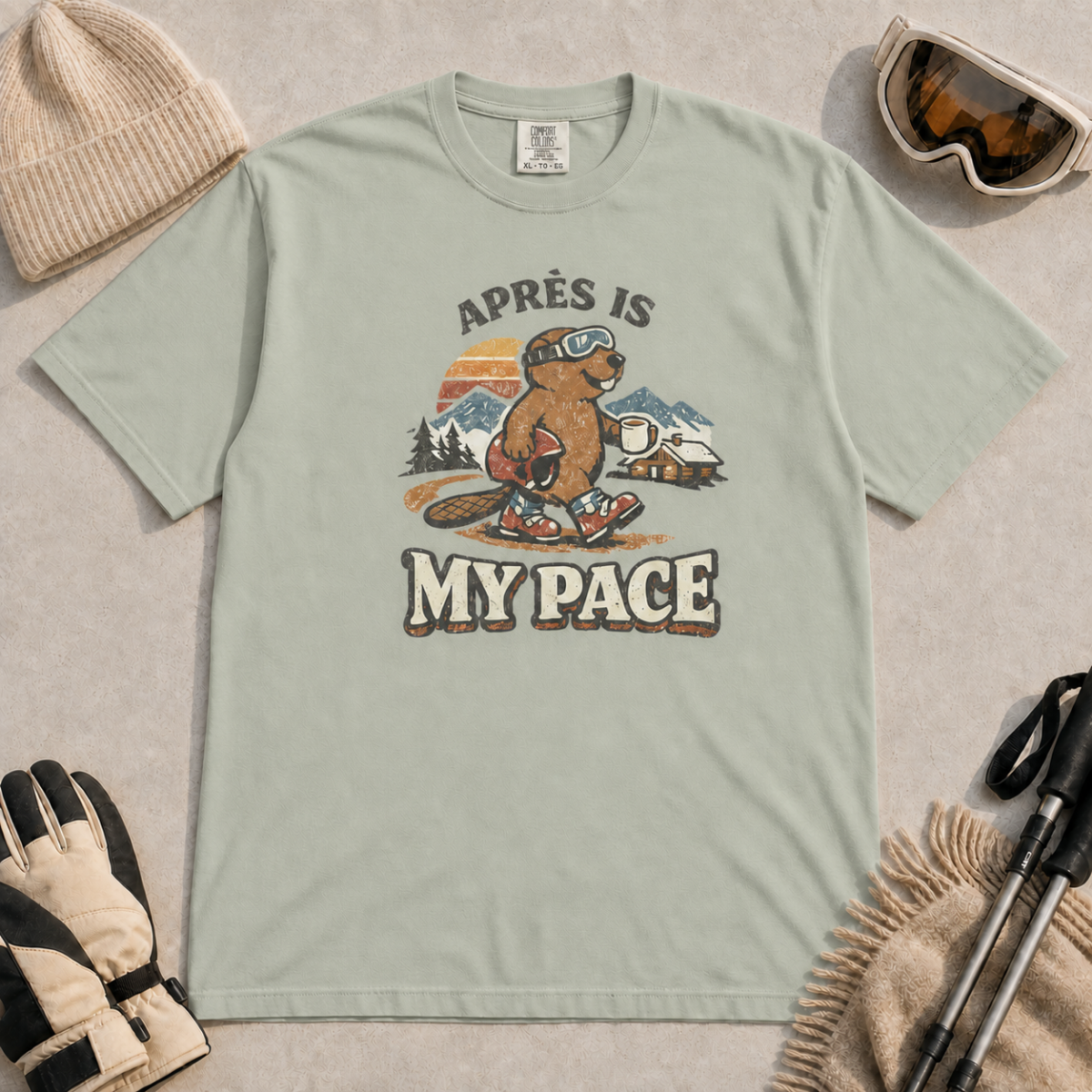 Bay t-shirt featuring an “Après Is My Pace” design with a beaver wearing ski goggles and boots, carrying a helmet and hot drink against a retro mountain and chalet scene.