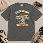 Pepper tee featuring a small mountain rodent holding a mug beside ski goggles, with a retro alpine sunset and “Elevation Without Expectation” text.