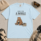 Chambray tee featuring a relaxed bear sitting on a bench with a warm drink beside ski boots and goggles, with the phrase “Here For A While.”