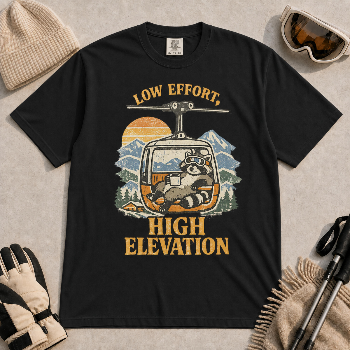 Black t-shirt featuring a vintage raccoon wearing ski goggles and lounging in a gondola with a mug, with the phrase “Low Effort, High Elevation.”