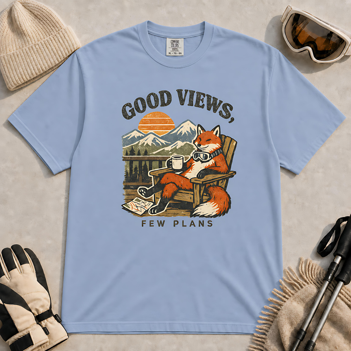Washed denim tee featuring a relaxed fox lounging in a wooden chair with ski goggles and a mug, framed by mountain scenery and the phrase “Good Views, Few Plans.”