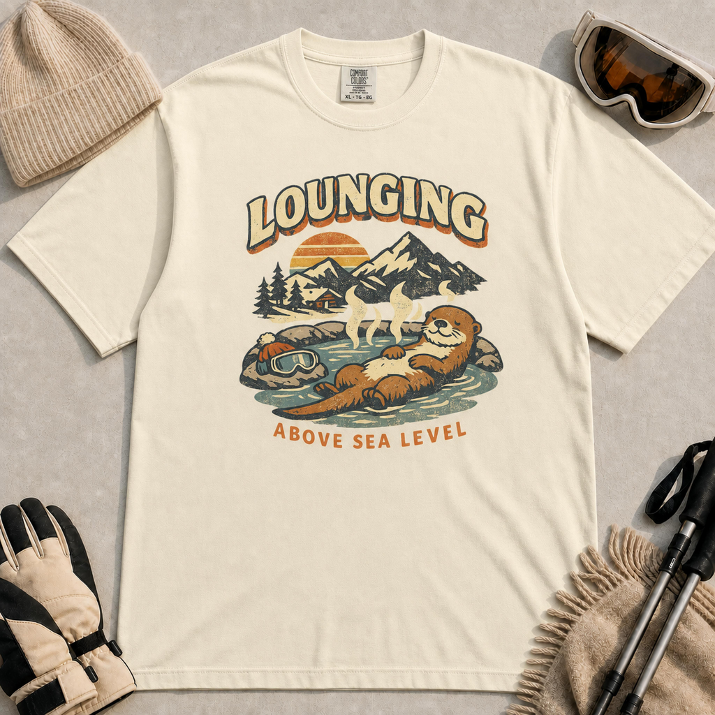 Ivory t-shirt featuring a vintage otter lounging in a mountain hot spring beside ski goggles, with the phrase “Lounging Above Sea Level.”
