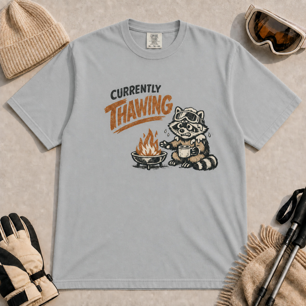 Granite tee featuring a shivering raccoon warming beside a campfire with a mug and the text “Currently Thawing.”