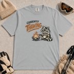 Granite tee featuring a shivering raccoon warming beside a campfire with a mug and the text “Currently Thawing.”
